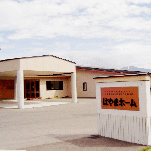 介護施設の建物外観。平屋造りで「やまぼホーム」と表示された看板が見える。